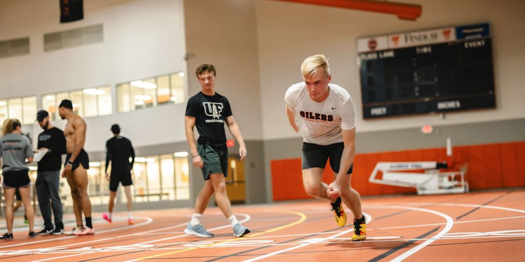 Two male athletes in a indoor facility, one is preparing to sprint.